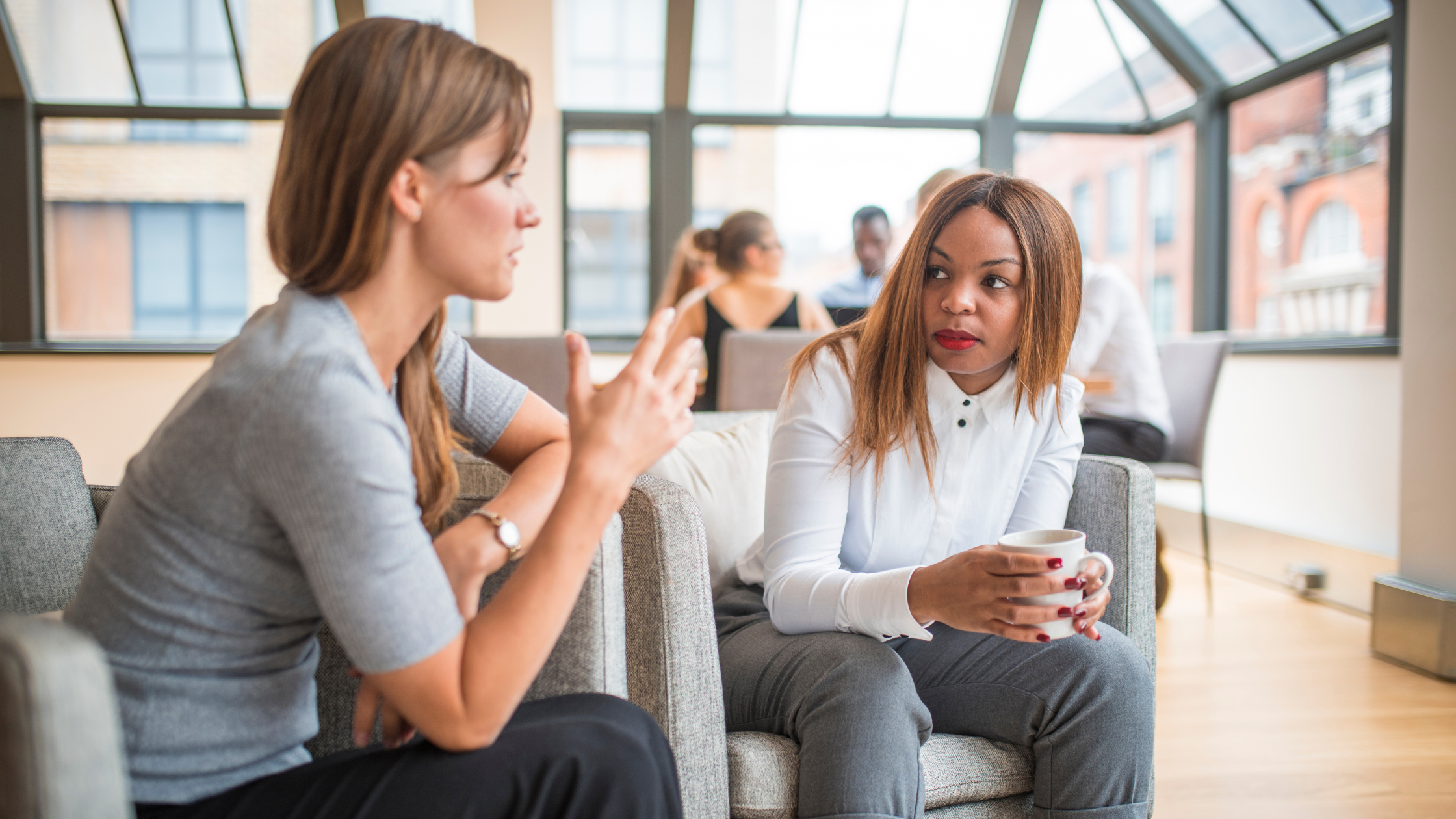 Convos Two people are sitting on a couch in a bright, modern room with large windows. One person is gesturing with their hands while speaking, and the other person is holding a cup and listening attentively. In the background, there are more people sitting at tables and engaging in conversation.