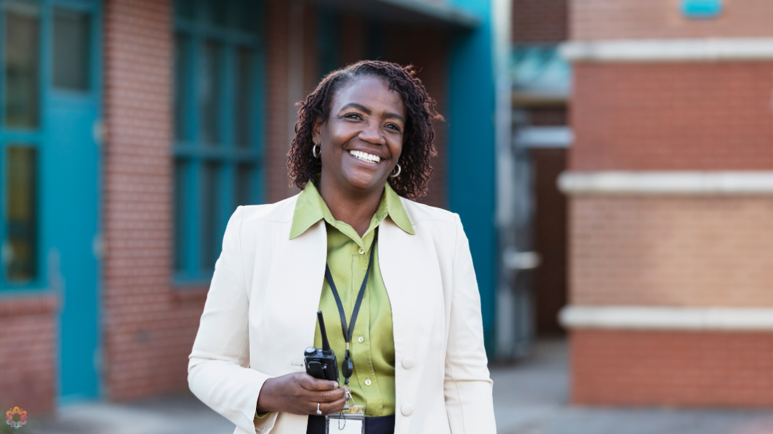 Principal of a school A person is standing outdoors in front of a building. They are wearing a light-colored blazer over a green shirt and holding a walkie-talkie in their right hand. The background includes blue-framed windows and brick walls.