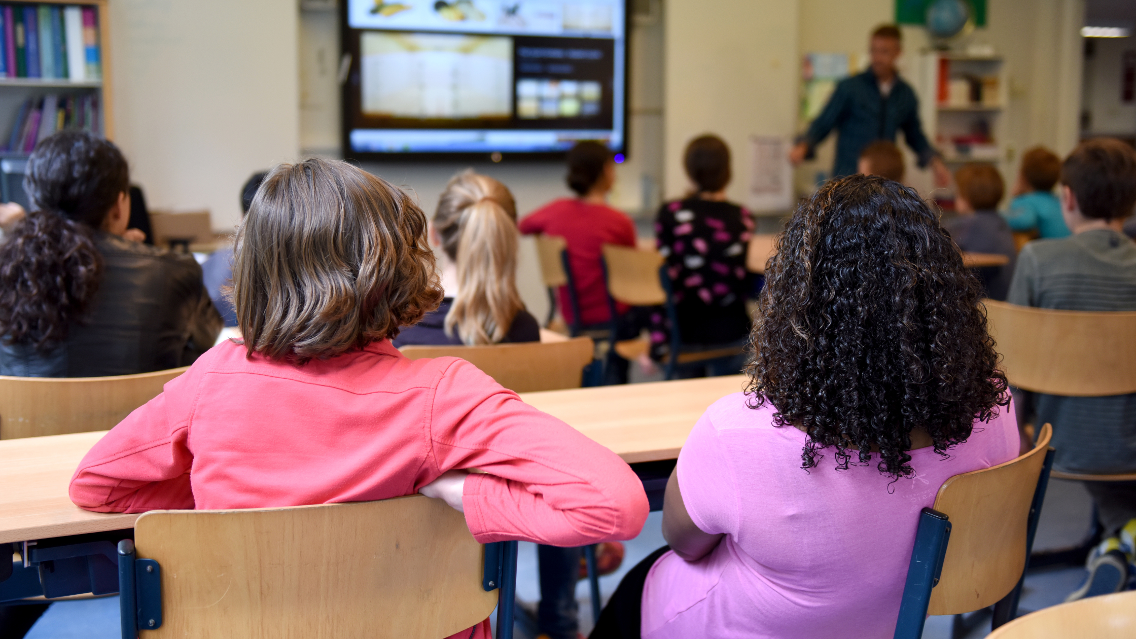 Students in the classroom A classroom scene with students seated at desks facing a teacher who is standing near a large screen displaying educational content. The students are of various ages and are attentively looking towards the front of the room. The classroom has bookshelves and educational materials visible in the background.