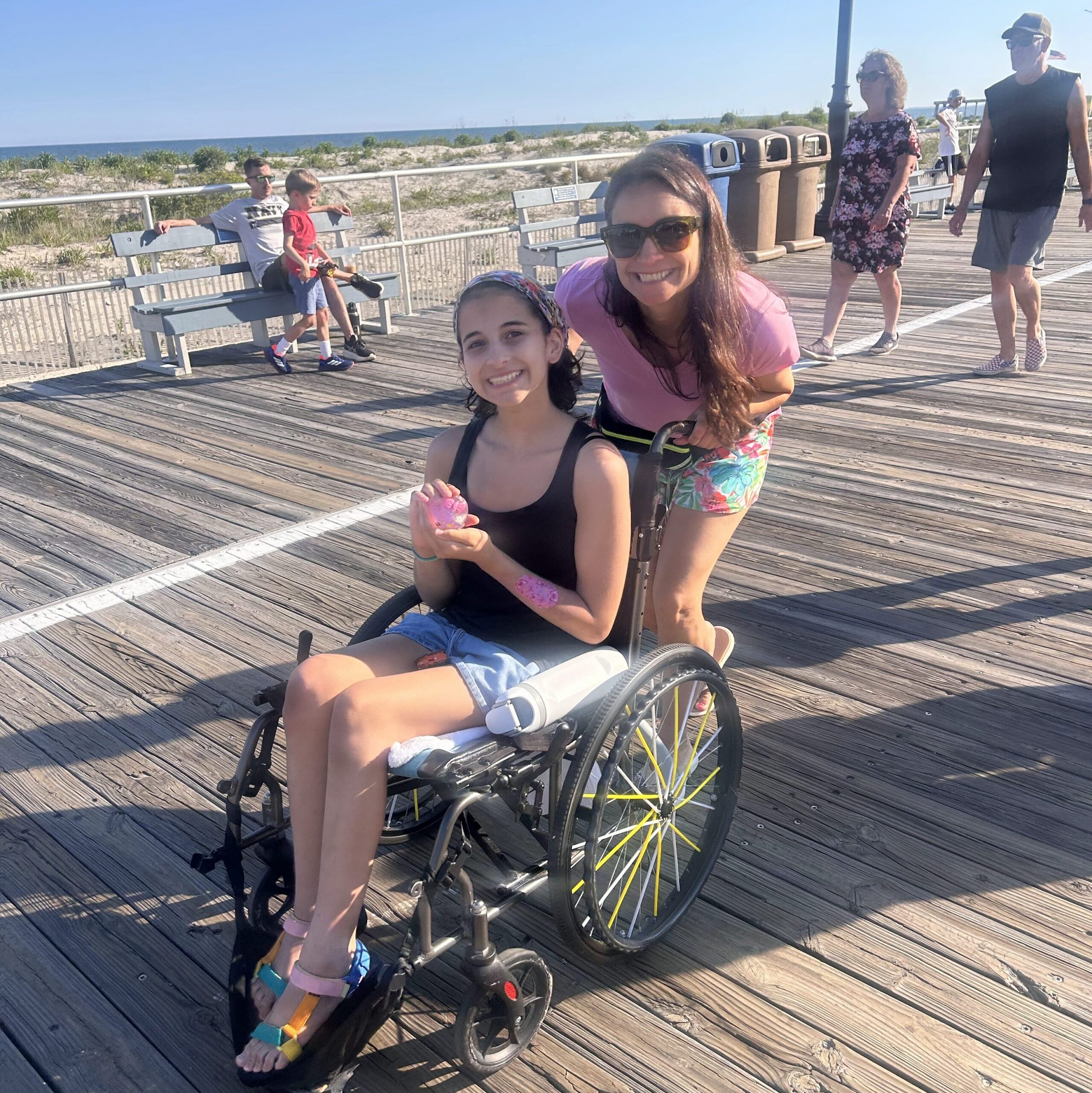 Jenna Rufo standing behind Emma Rufo in her wheel chair on a beach boardwalk