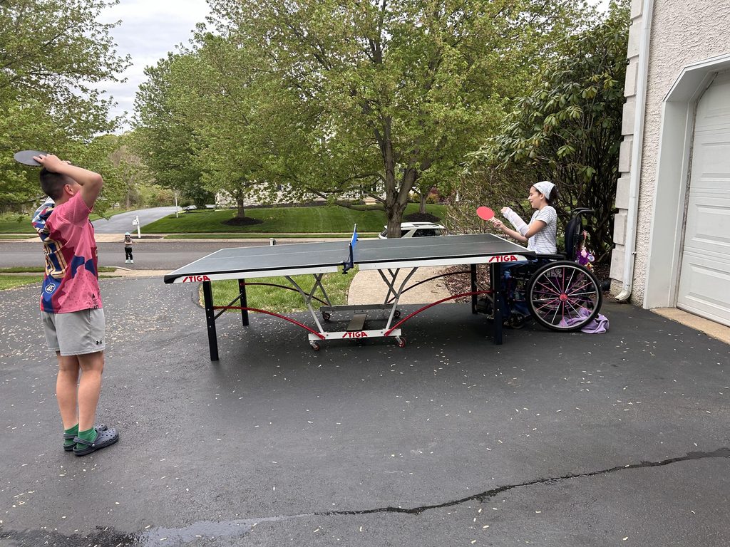 Emma Rufo in her wheel chair, wearing a white shirt and bandana playing ping pong on a driveway against a person in a pink shirt.